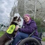 A woman in a wheelchair with a dog in front of a radio telescope.